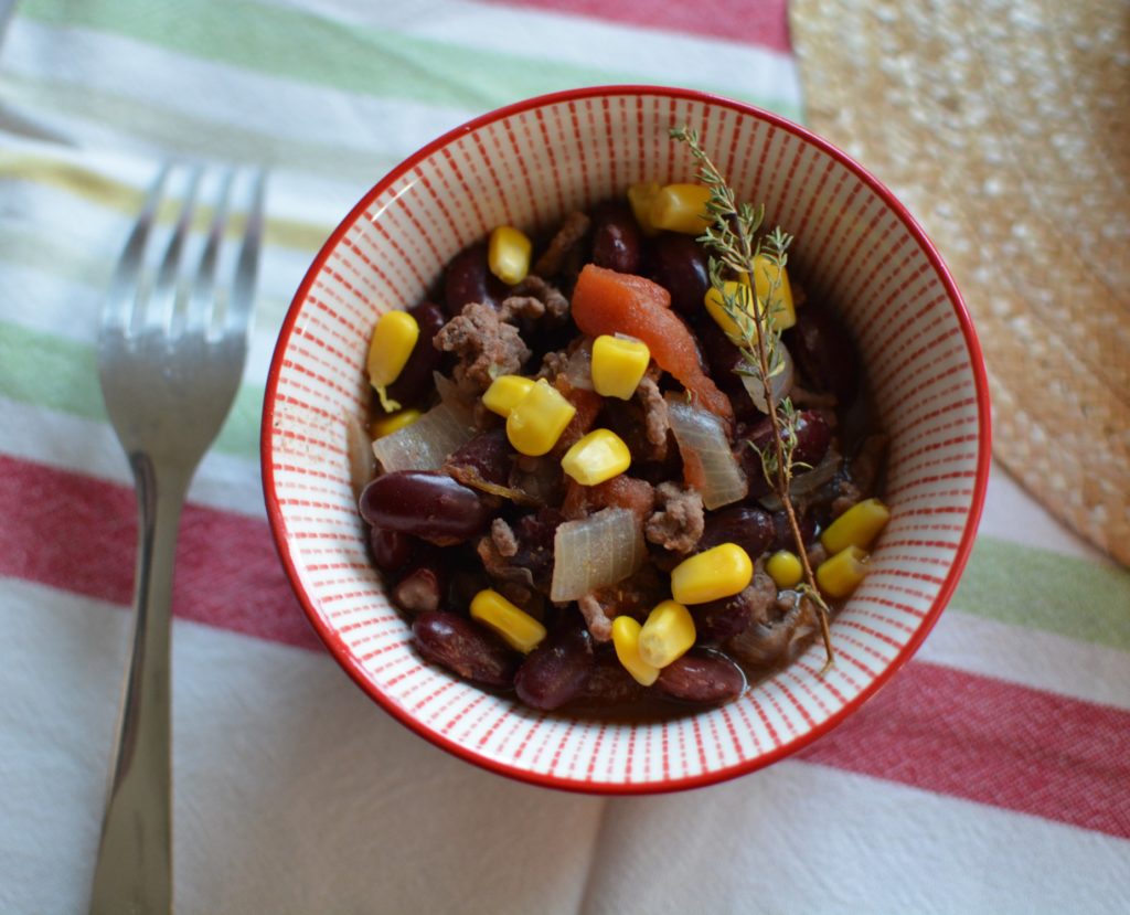 Bol rouge contenant des haricots rouges, du maïs, de la viande, des oignons et du thym. Posé sur une serviette colorée à côté d'une fourchette et d'un chapeau de paille