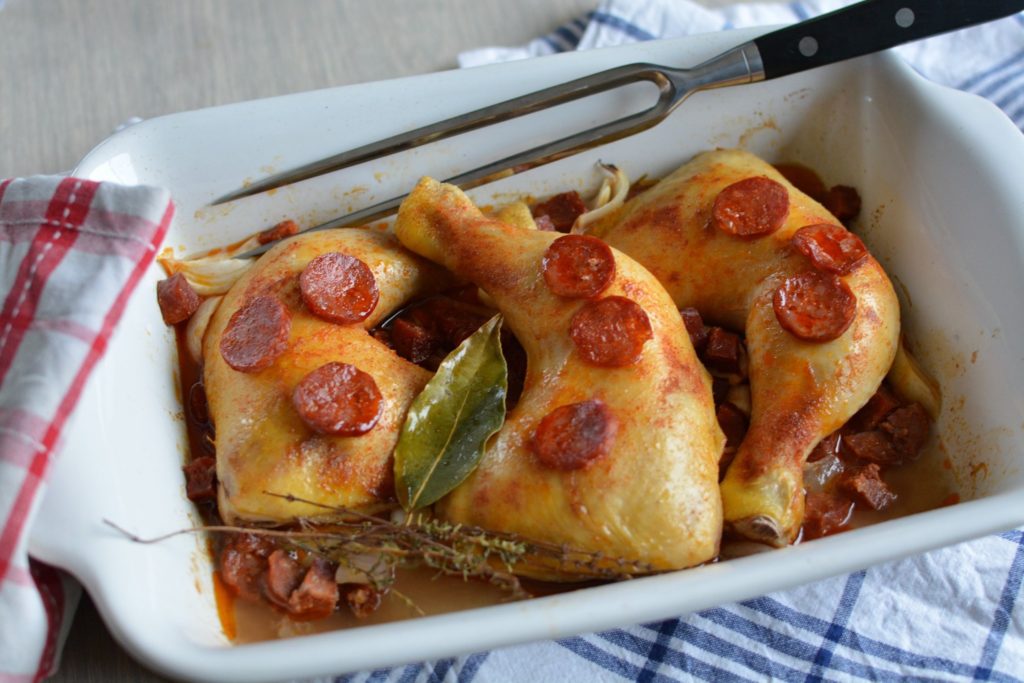 3 cuisses de poulet cuits dans un plat blanc avec des tranches de chorizo parsemés dessus, une fourche de cuisine posée sur le plat et un torchon posé à côté.