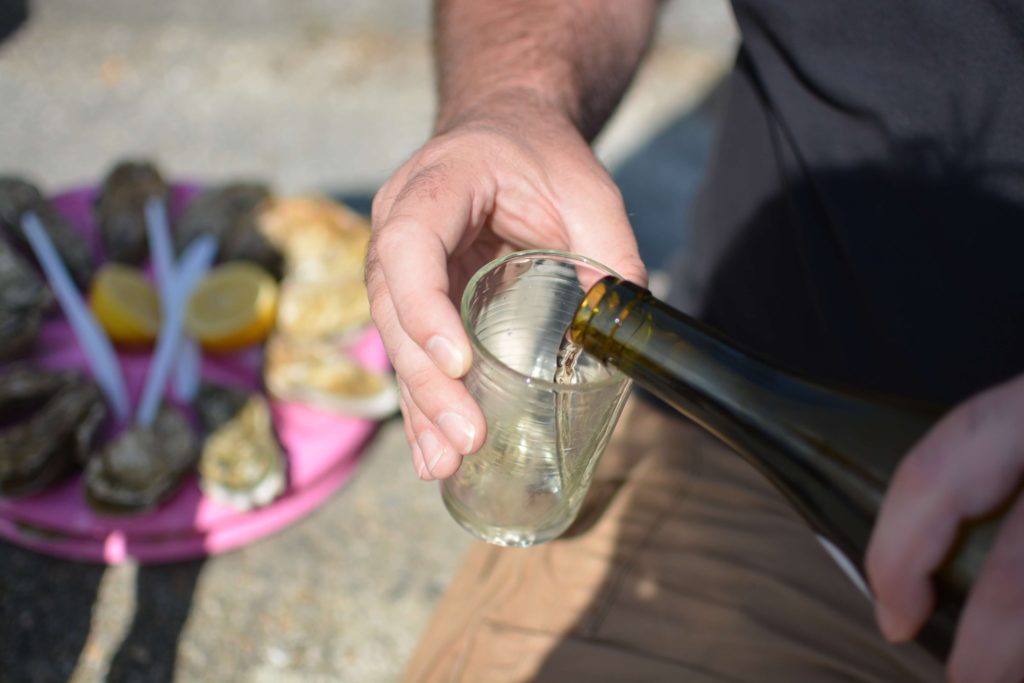 Mains d'homme en train de servir un verre de vin blanc à côté d'un plateau d'huîtres posé au sol