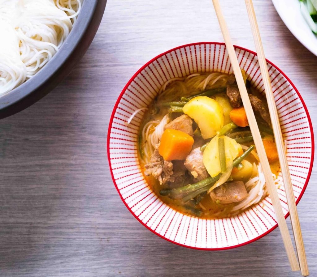 Bol rouge contenant un bouillon de curry rouge avec des légumes, des nouilles et de la viande. 