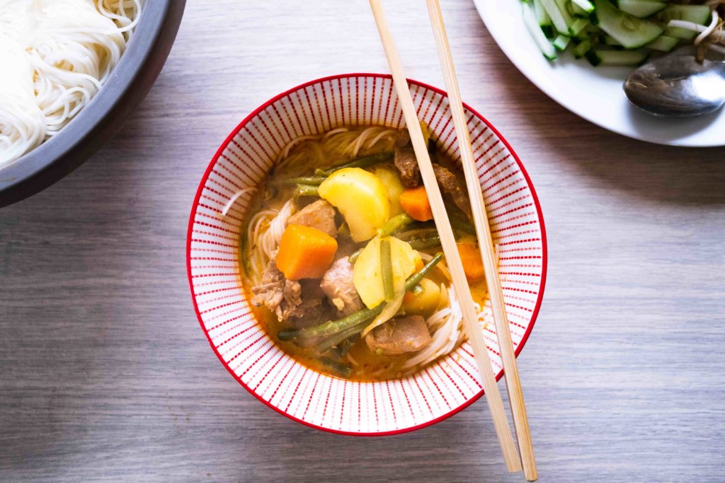 Bol rouge contenant un bouillon de curry rouge avec des morceaux de pommes de terre, des carottes, des nouilles blanches, de la viande et une paire de baguettes posées dessus