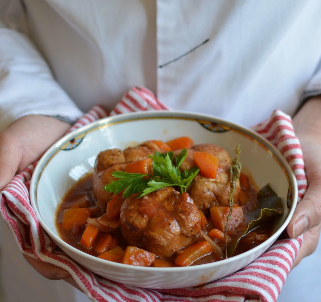 Mains tenant une serviette rouge sur laquelle est posée une assiette de paupiettes à la sauce tomate, avec des morceaux de carottes, une branche de thym, une feuille de laurier et du persil pour décorer