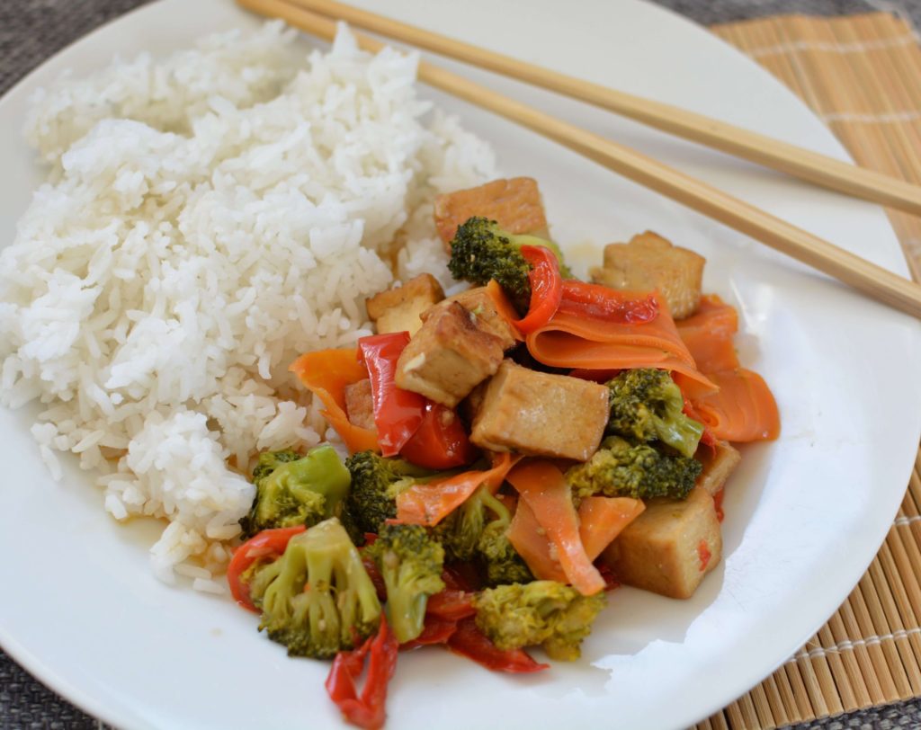 Assiette blanche contenant du riz, et des morceaux de tofu avec des carottes en lamelles, du brocoli et des morceaux de poivron. Le tout posé sur un set en bambou avec une paire de baguettes