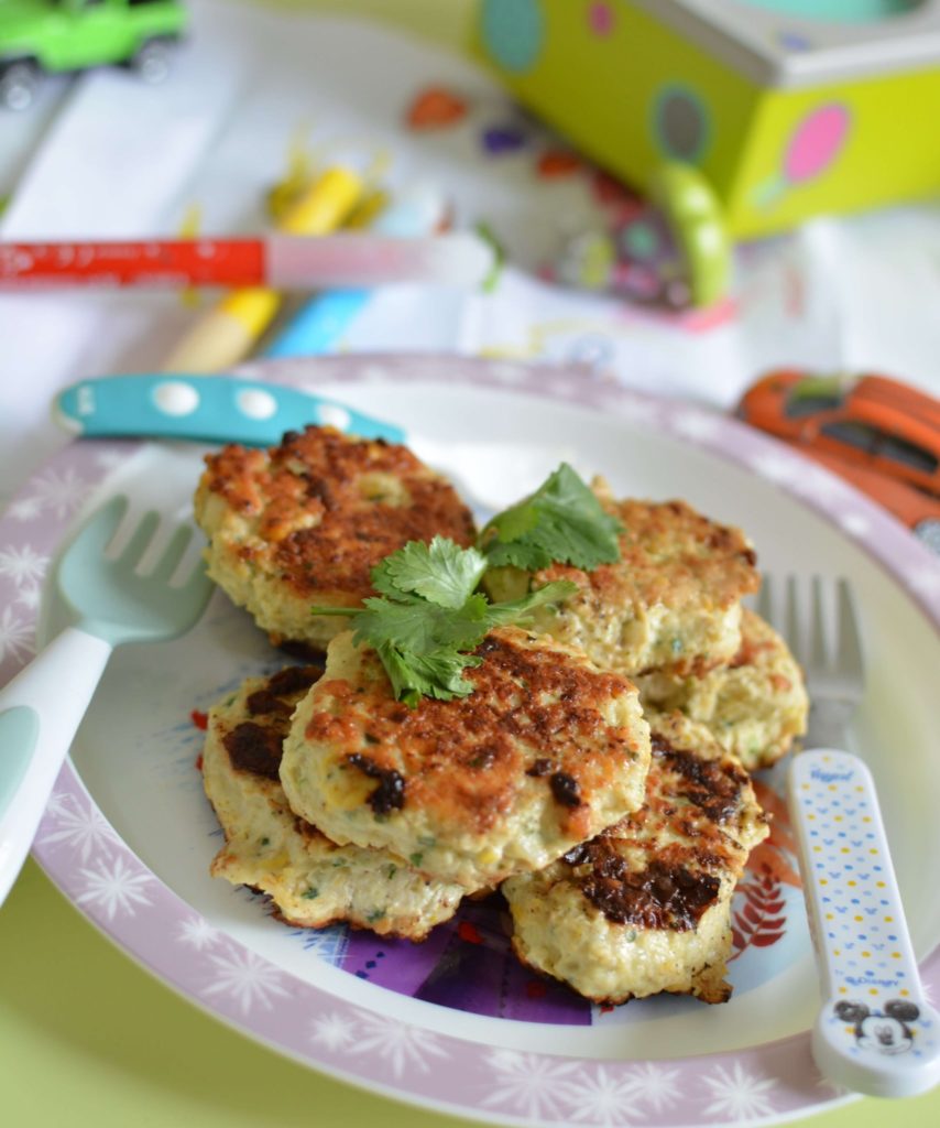 Croquettes de poulet posées dans une assiette décorée pour enfants, avec des fourchettes pour enfants de couleurs, et un brin de coriandre. Des crayons, des jouets et des dessins d'enfants en fond.