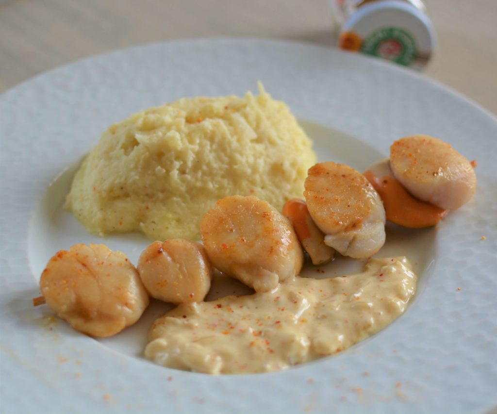 Assiette blanche avec une brochette de 5 noix de saint jacques, une sauce à la crème et une purée. En fond, un pot de piment d'Espelette couché