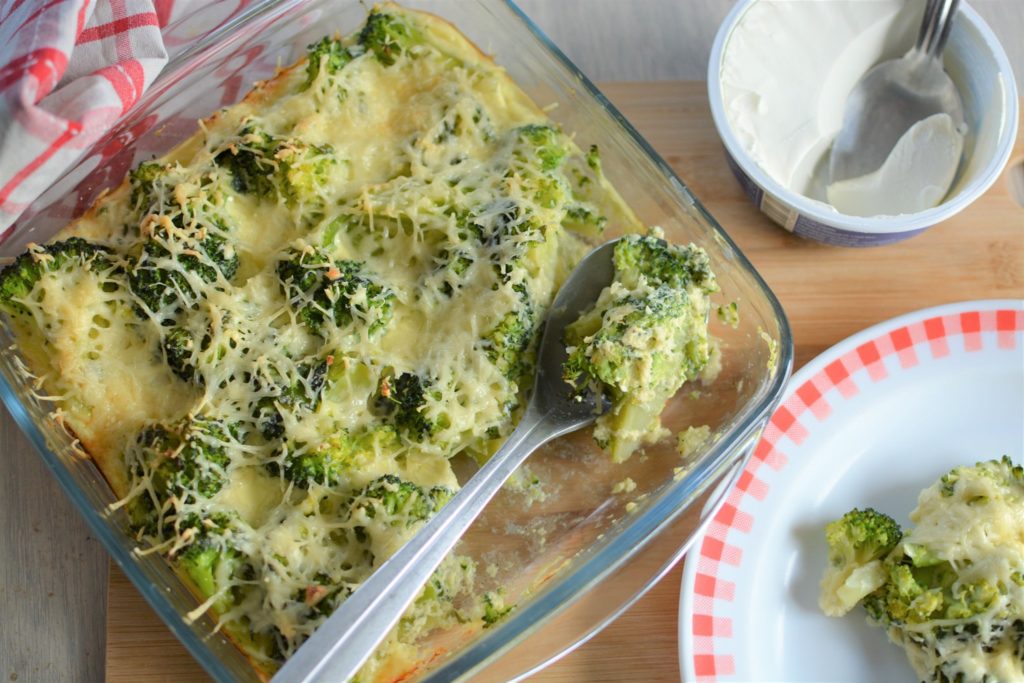 Plat contenant des brocoli et du fromage gratiné avec une cuillère de brocoli, posé sur une planche en bois à côté d'une assiette de brocoli et un pot de crème