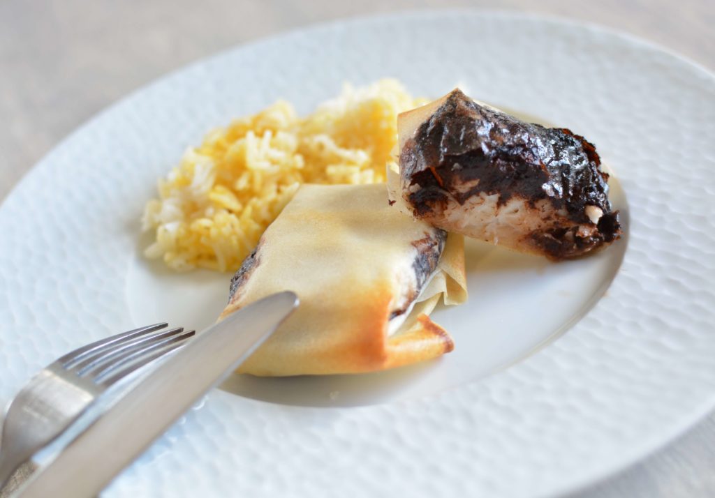 Assiette blanche contenant du riz, un feuilleté entier et un feuilleté coupé contenant du poisson blanc et de la tapenade noire. Posés sur l'assiette, une fourchette et un couteau