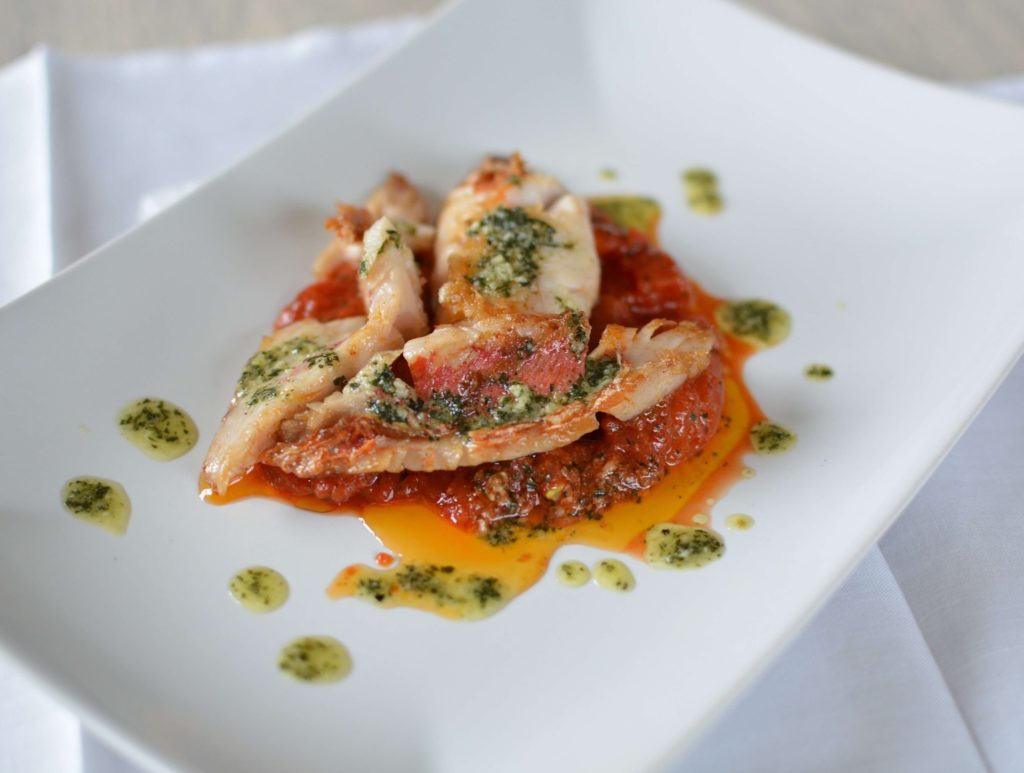 3 filets de poissons à la peau rouge posés sur un lit de sauce tomate avec autour des gouttes de sauce vertes, le tout sur une assiette rectangulaire