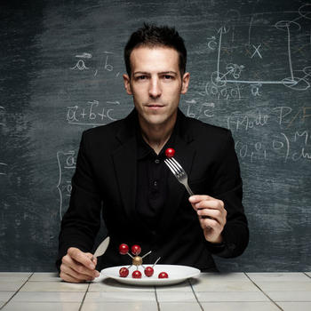 Homme assis devant un tableau avec des formules écrites à la craie, face à une assiette avec un modèle de molécule chimique et portant à sa bouche un morceau de molécule